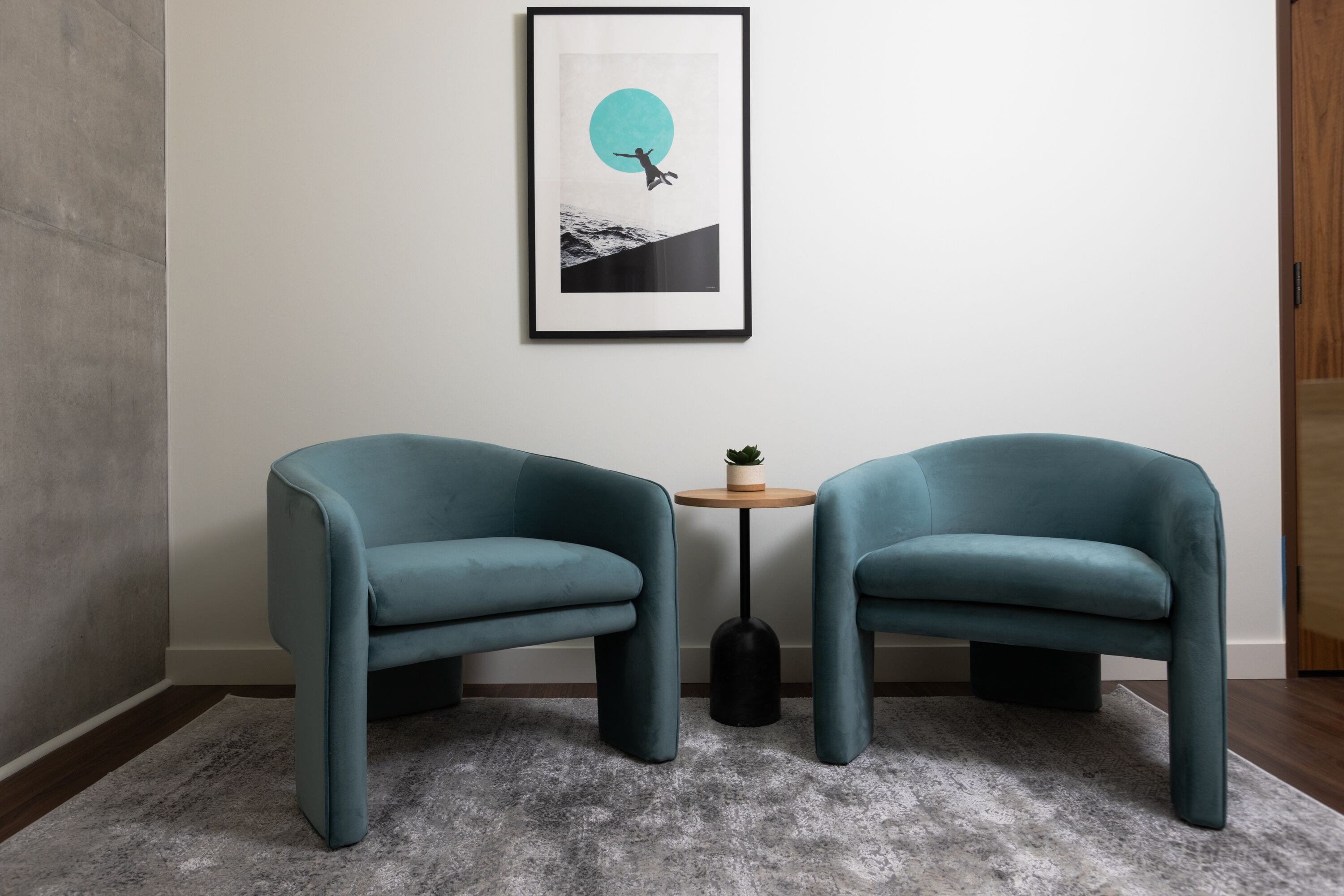 Two teal armchairs, a framed artwork, and a small wooden table with a potted plant.