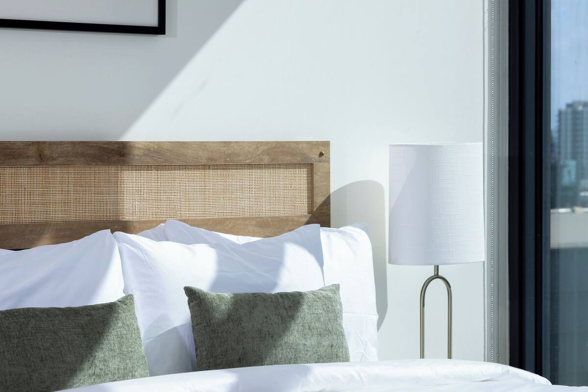 Een net opgemaakt bed met witte lakens en groene kussens, een bedlamp en een lijst met een afbeelding aan de muur.