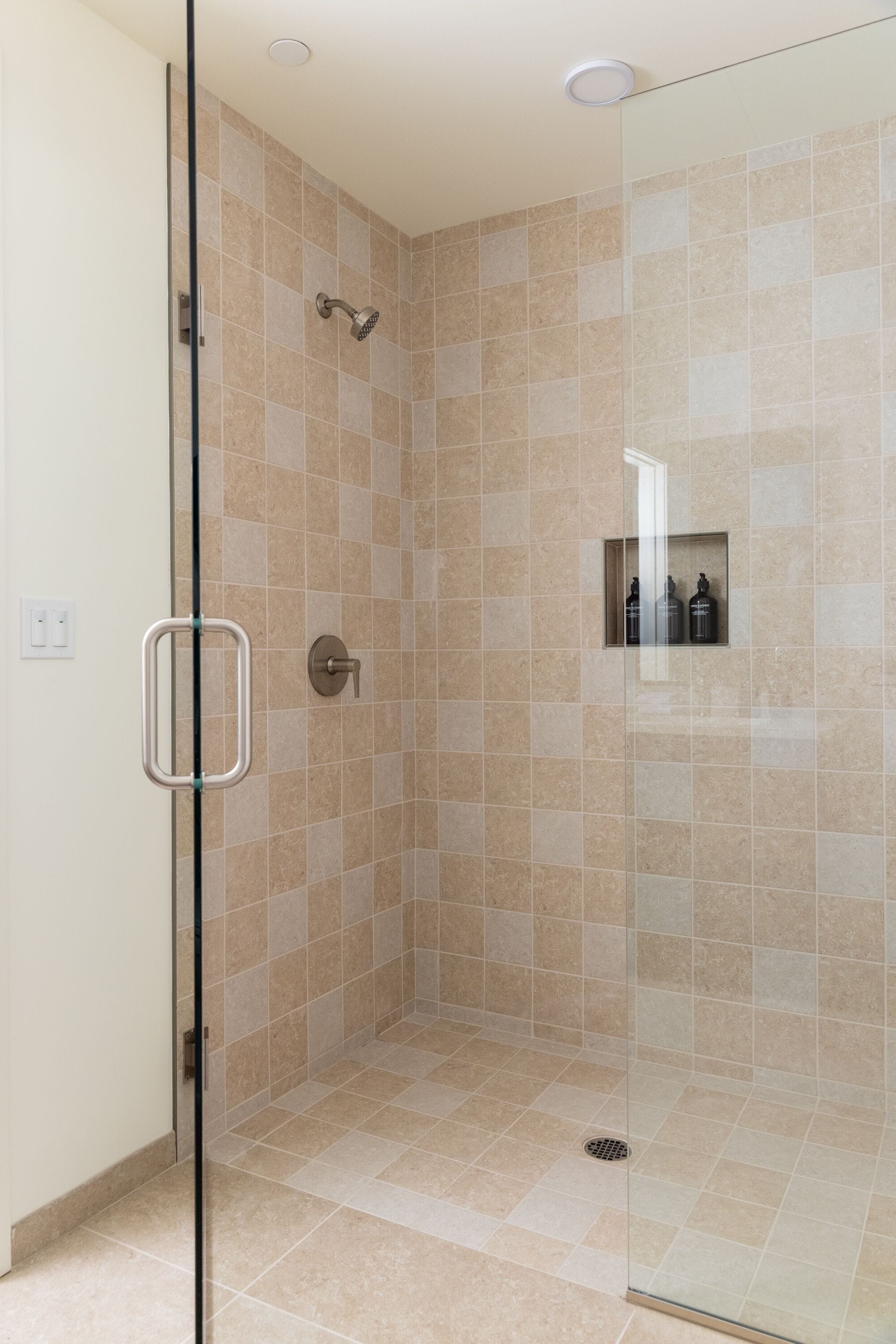 Um chuveiro com paredes de vidro, azulejos bege, um chuveiro visível e uma pequena prateleira com frascos.