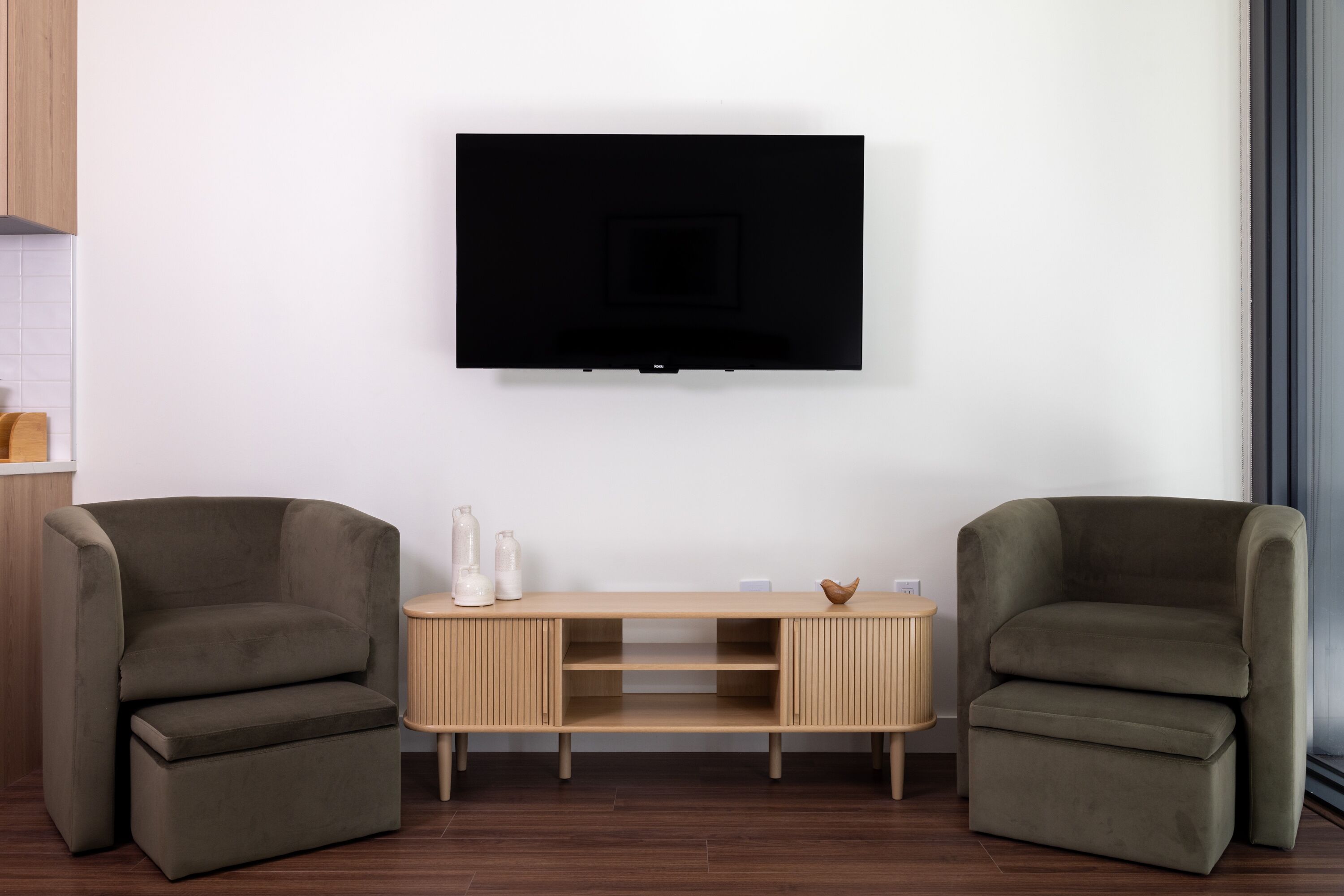 Ein modernes Wohnzimmer mit flachbildfernseher an der Wand, zwei Sesseln, einem Holzkonsolentisch mit Dekoration und Holzboden.