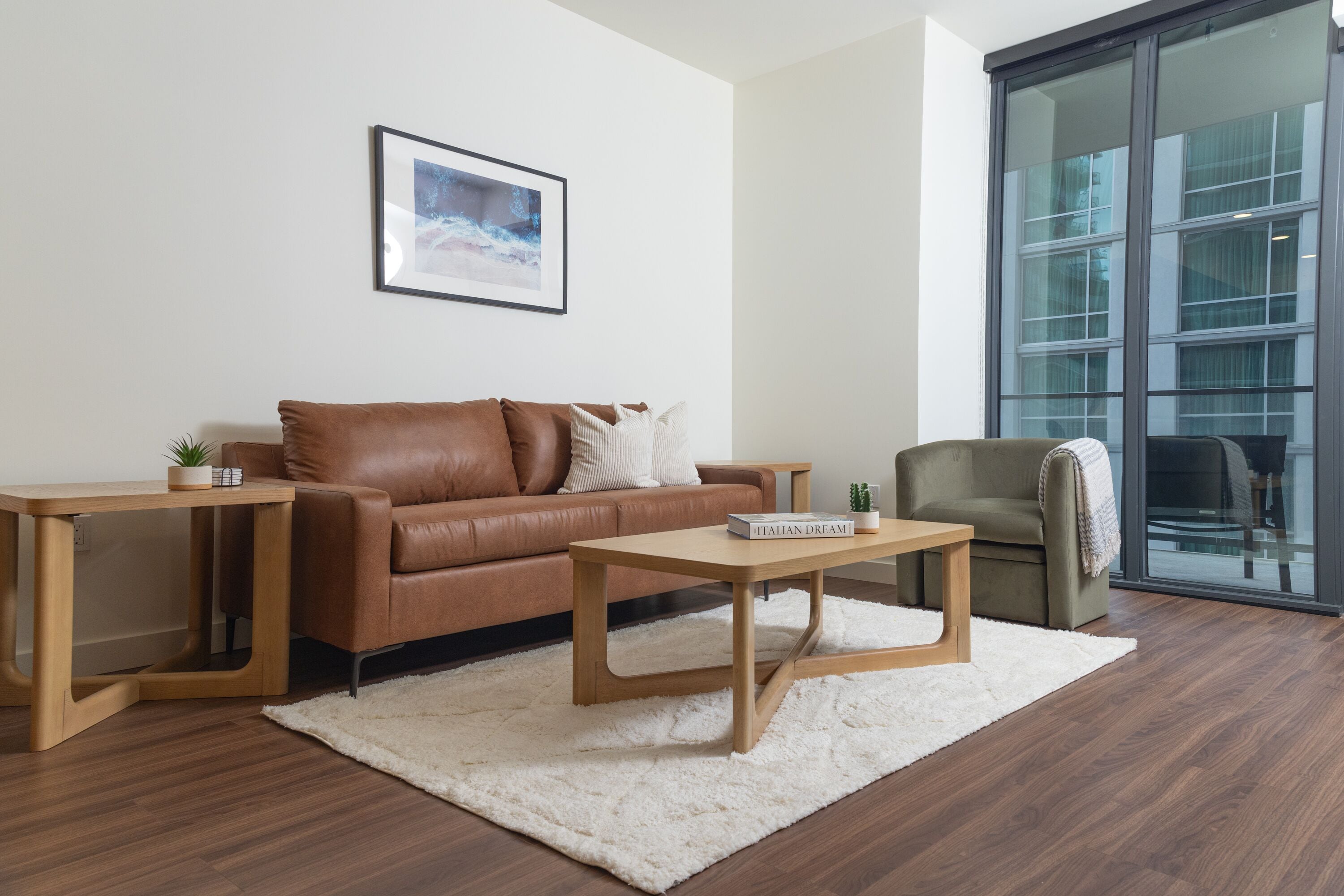 Ein modernes Wohnzimmer mit einer braunen Ledersofa, einem Holztisch und einem grünen Sessel. An der Wand hängt ein gerahmtes Bild und ein großes Fenster bietet Blick auf Gebäude.