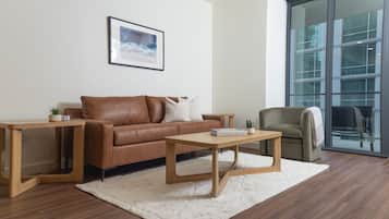Una sala moderna con un sofá de cuero marrón, una mesa de centro de madera y un sillón verde. Hay un cuadro enmarcado en la pared y un ventanal con vistas a edificios.