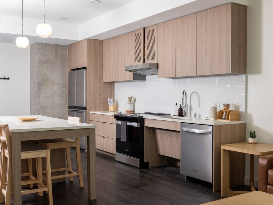 A modern kitchen with wooden cabinets, a stainless steel refrigerator, a black oven, and a dishwasher. There is a wooden dining table with two chairs and a small wooden side table with a plant.