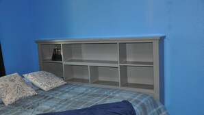 Un dormitorio con pared azul, cama con colcha azul y blanca a cuadros, dos almohadas estampadas y cabecero blanco con varios estantes.
