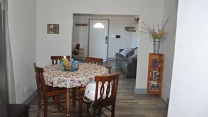 Un comedor con una mesa redonda, sillas y mantel con estampado floral. Se observa una estantería con libros y un jarrón con flores.