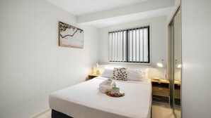 Habitación de hotel ordenada con una cama, una mesita de noche con lámpara, una ventana con persianas, una pequeña mesita de noche con una botella y un cuadro en la pared.