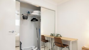 Ein Zimmer mit Schreibtisch, zwei Stühlen, einem Spiegel, einem Kleiderschrank mit Regalen und einem Wäschekorb.