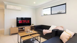 Un salon moderne avec un téléviseur à écran plat affichant le logo « NETFLIX », un canapé, une table basse et une fenêtre munie d’un moustiquaire.