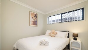 Un lit bien fait, avec du linge de lit blanc et une serviette enroulée, une petite table de chevet avec une lampe, une fenêtre avec des stores et un tableau encadré au mur.
