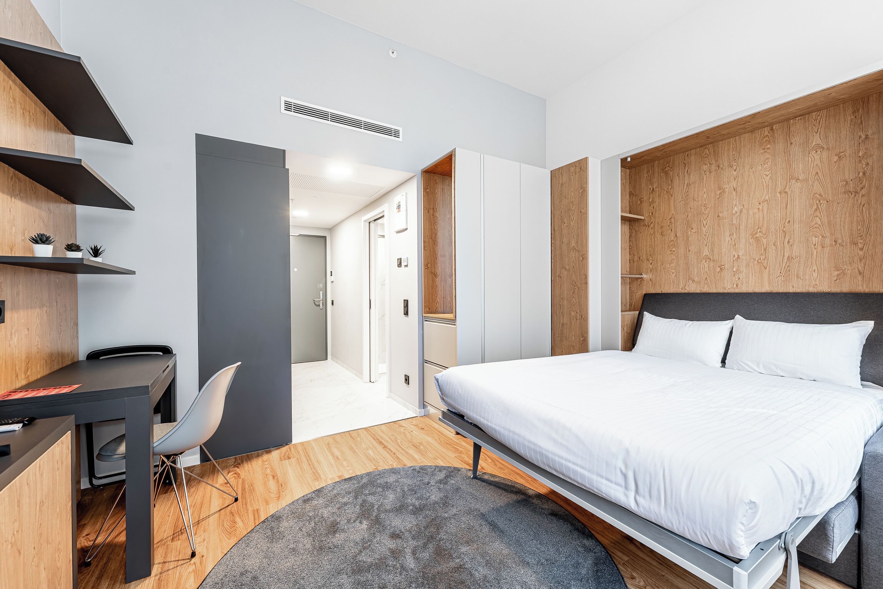 Ein modernes Schlafzimmer mit einem Bett, einem Schreibtisch und einem Stuhl. Ein Kleiderschrank und eine Tür zu einem anderen Raum sind ebenfalls vorhanden.