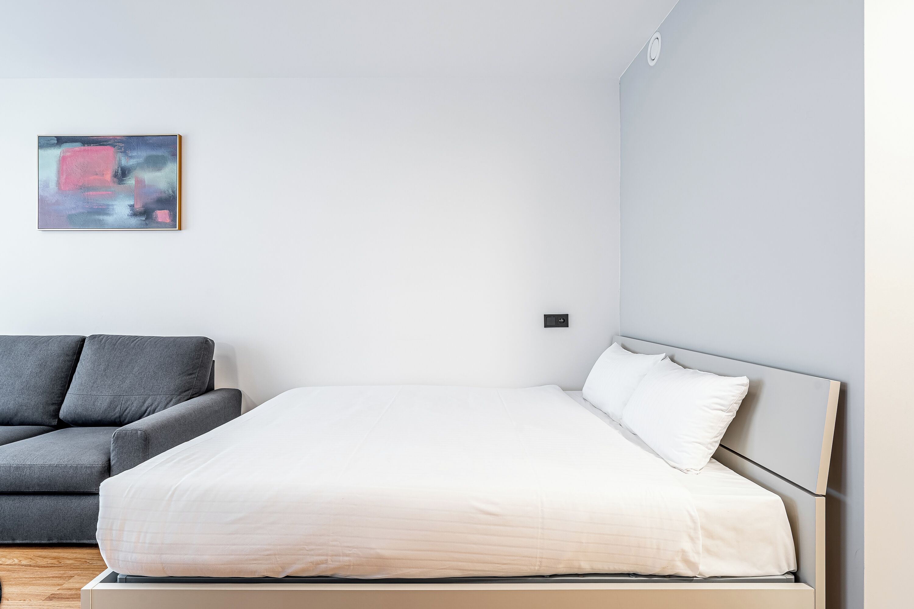 A modern bedroom with a bed, a gray sofa, and a minimalist abstract painting on the wall.