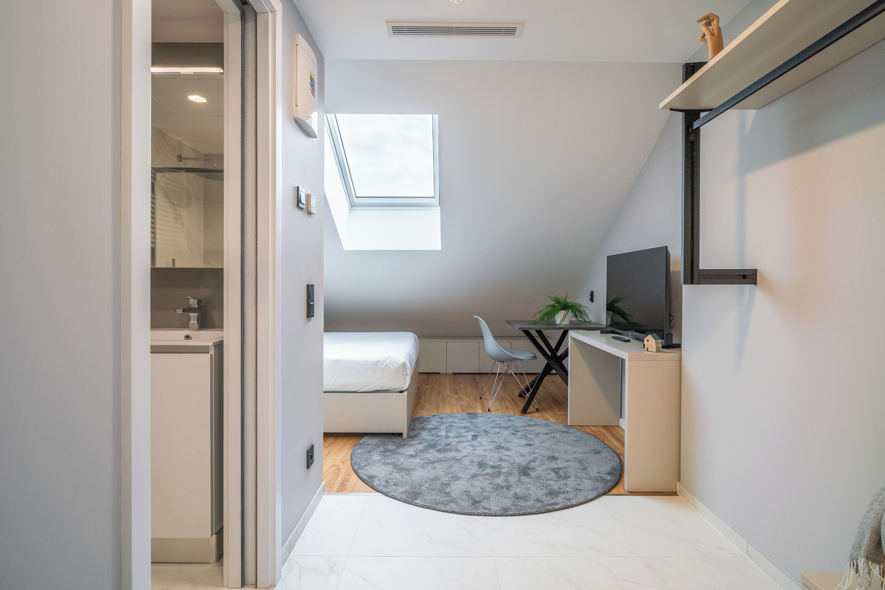 A compact bedroom with a bed, a desk, a chair, a small table with a plant, and a window.