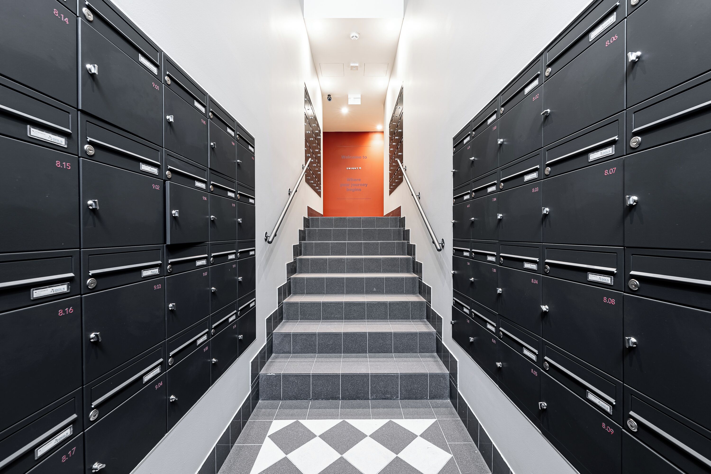 A set of black metal lockers with numbered labels, arranged in two rows on either side of a staircase leading to an orange door.