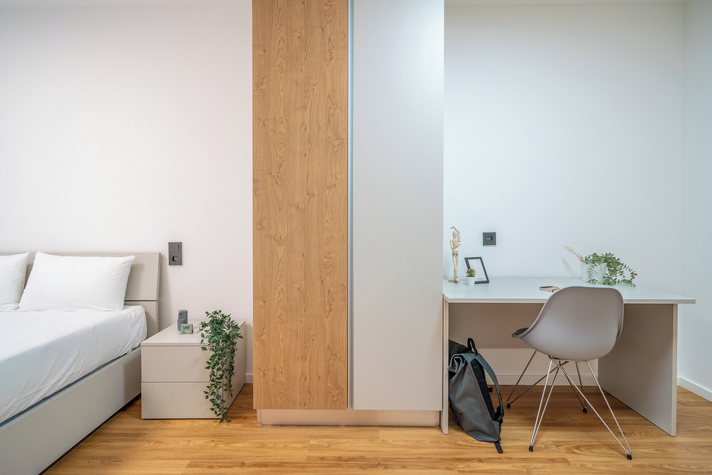 A bedroom with a bed, a bedside table with a plant, a desk with a chair, and a wall-mounted light.