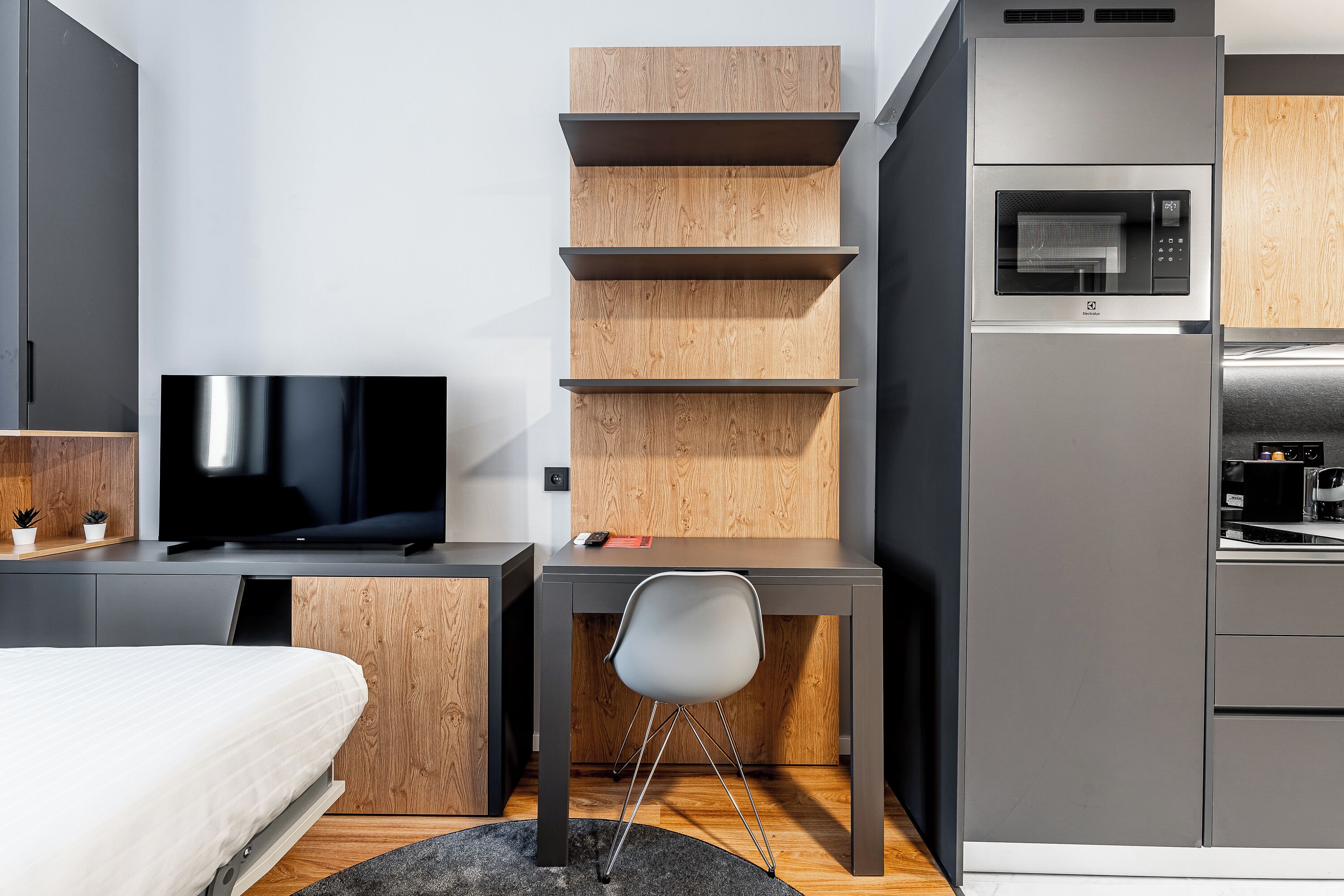 A compact hotel room with a bed, a TV, a desk with a chair, a microwave, and a small kitchenette.