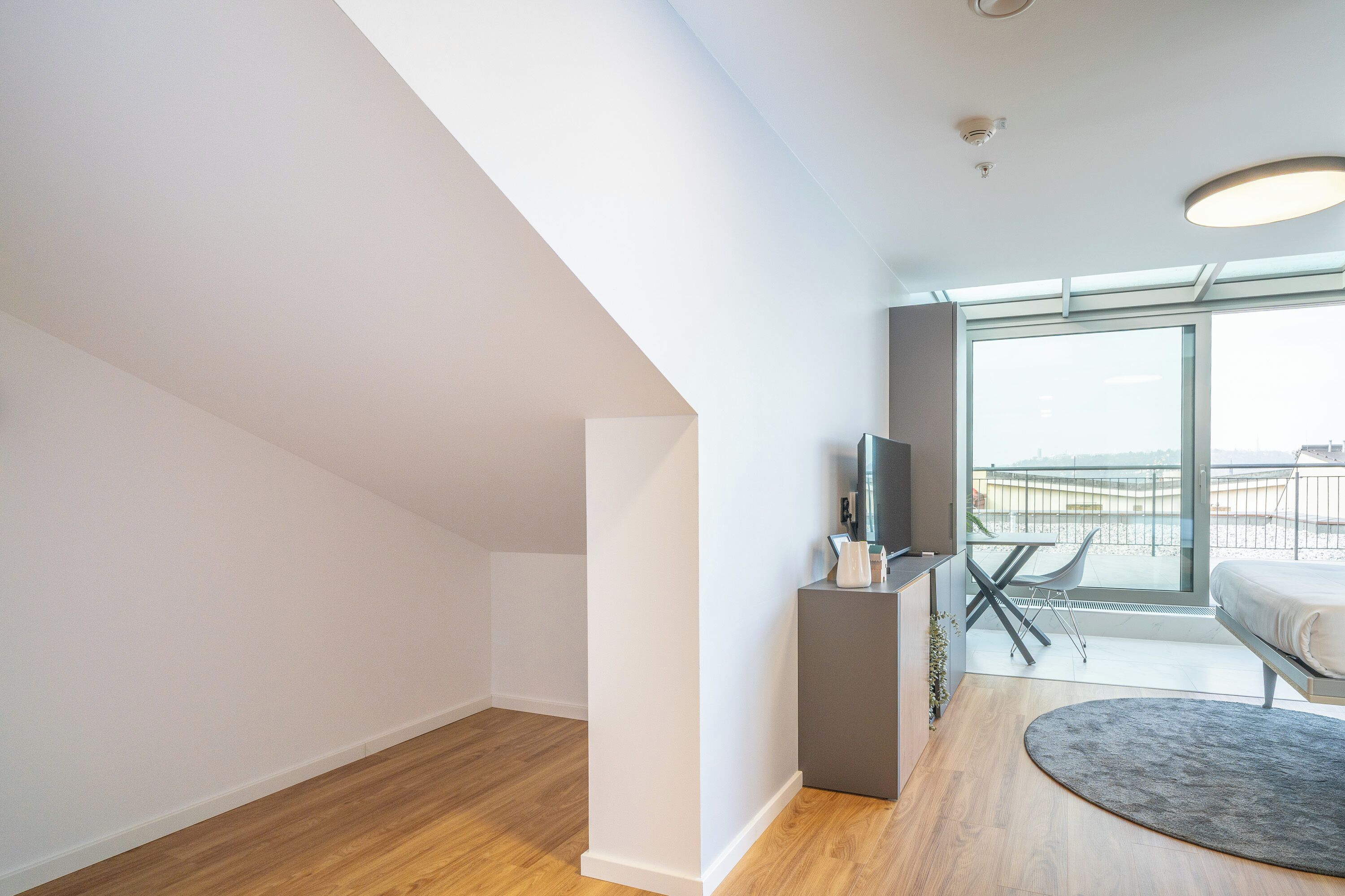 A modern bedroom with a large window, a bed, a TV, and a small seating area.