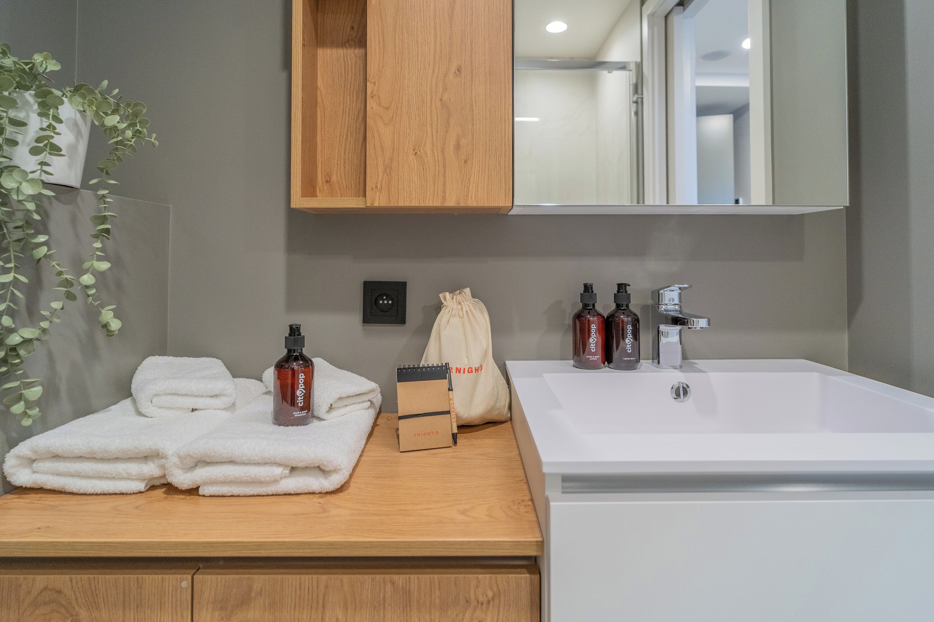 A bathroom with a wooden vanity, a white sink, and a mirror above it. There are towels, a small box, and two bottles on the vanity.