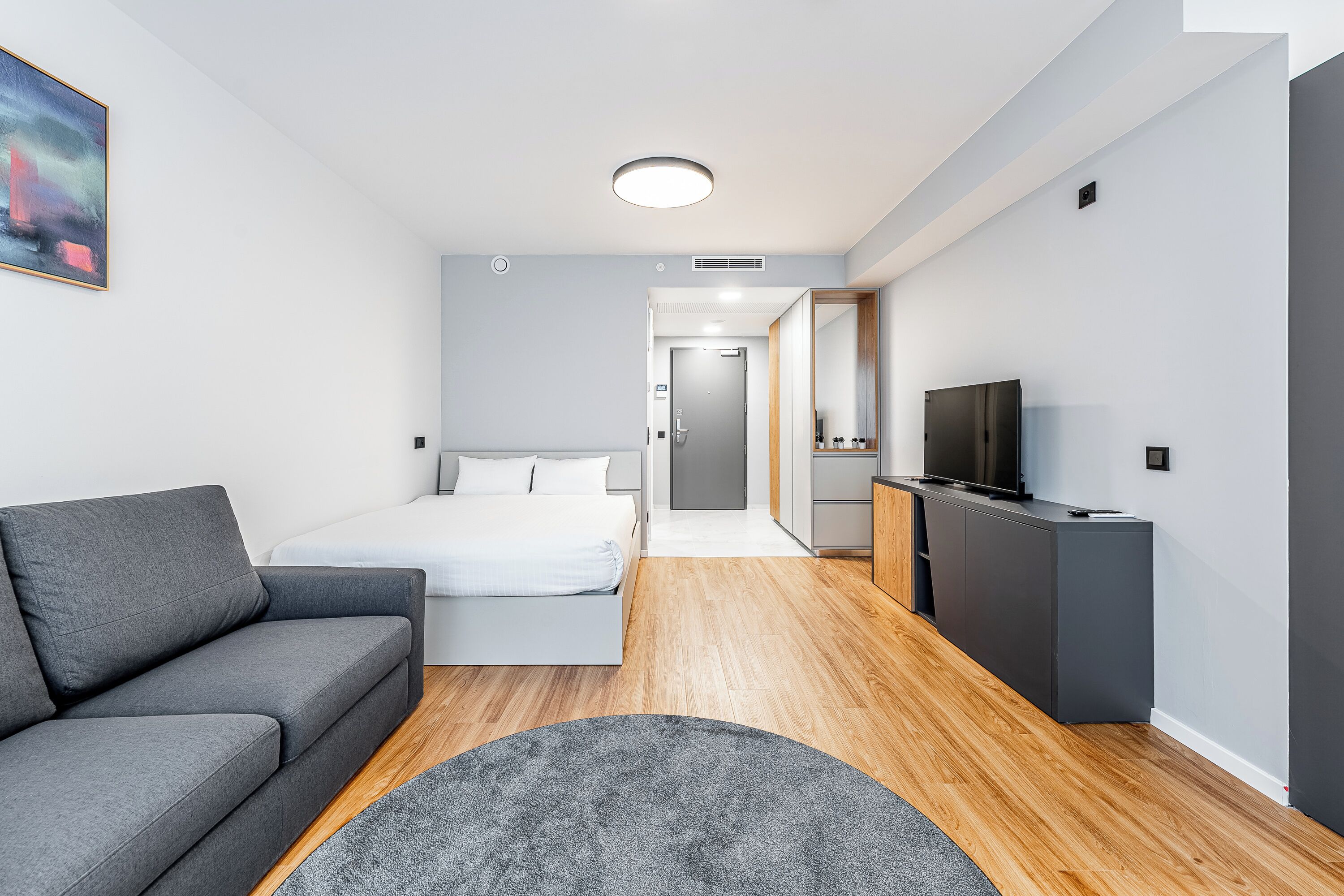 A modern hotel room with a grey sofa, a bed, a flat-screen TV, and a wooden floor.