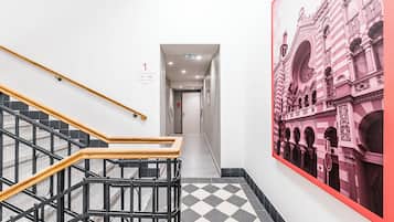 Un corridoio con pavimento a scacchi, una scala con ringhiere nere e gradini in legno, e un grande murale raffigurante un edificio.