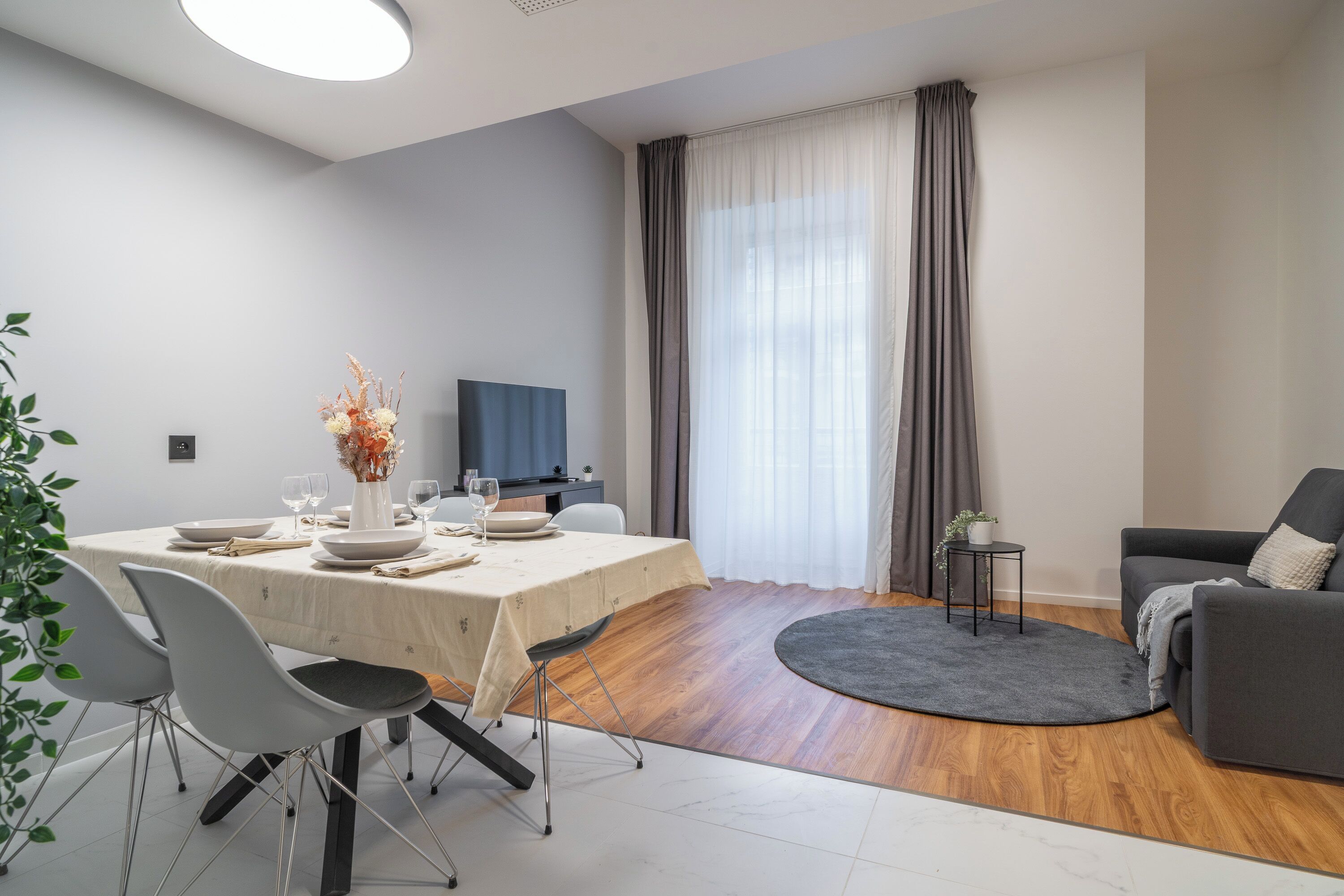 En moderne stue med spiseplads, en grå sofa, et lille sofabord og et fjernsyn i baggrunden.