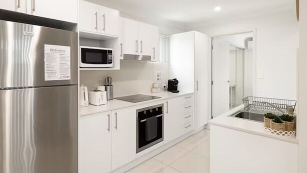 A modern kitchen with white cabinetry, stainless steel appliances, a microwave, a coffee maker, and a small sink with a dish rack.