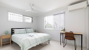Une chambre avec un lit, un bureau et une chaise. Il y a une fenêtre avec des stores, un ventilateur de plafond et un appareil de climatisation.