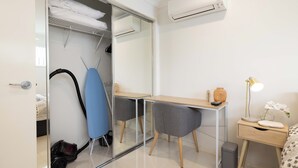 Une chambre moderne avec un placard en verre, un bureau en bois, une chaise, une table de chevet avec une lampe et un climatiseur.