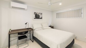 Une chambre à coucher avec un lit, un bureau et une chaise, un ventilateur de plafond, un climatiseur, une fenêtre avec des stores et un tableau encadré au mur.