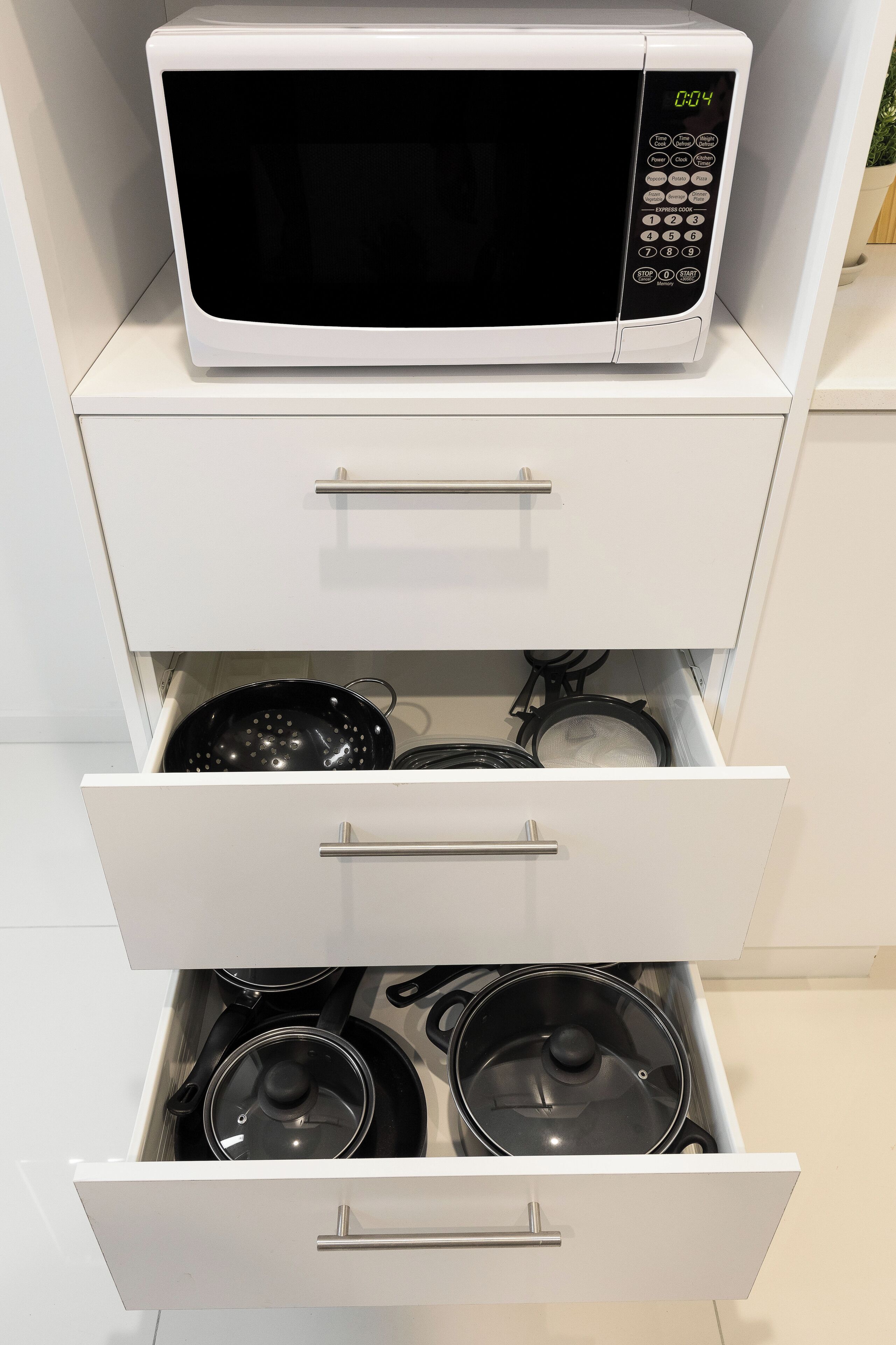 Un four à micro-ondes est posé sur une étagère d’un meuble blanc, et un tiroir situé en dessous contient une casserole et une poêle.