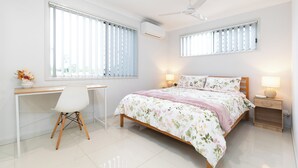 Un dormitorio con cama, escritorio, silla, mesitas de noche y ventanas.