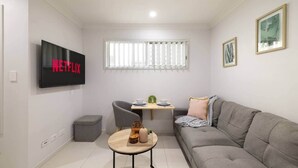 Una sala de estar moderna con un sofá gris, una mesita redonda pequeña, un sillón gris, un televisor montado en la pared y una ventana con persianas.