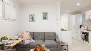 Una sala moderna con un sofá gris, una mesa de centro redonda con objetos decorativos, una zona de comedor con una mesa de madera y platos blancos, y una cocina con armarios blancos y una estufa.
