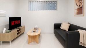 Een moderne woonkamer met een zwarte bank, een houten salontafel, een televisie waarop 'NETFLIX' staat, en een raam met zonwering.