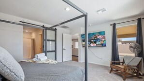 Ein Schlafzimmer mit Bett, Stuhl, Ventilator und einem durch eine offene Tür einsehbarer Badezimmer.