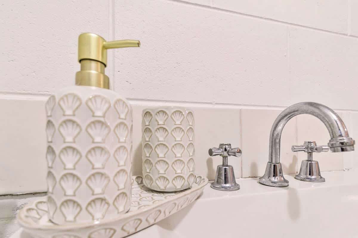 A bathroom with a white tiled wall, a white sink with a gold faucet, and a white soap dispenser with a gold top.