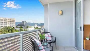 A balcony with two chairs, a small table with a plant, and a view of buildings and mountains.