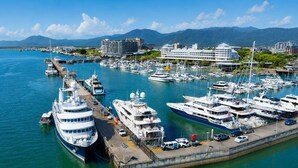 A marina with multiple yachts docked, a large building with a white facade, and mountains in the background.