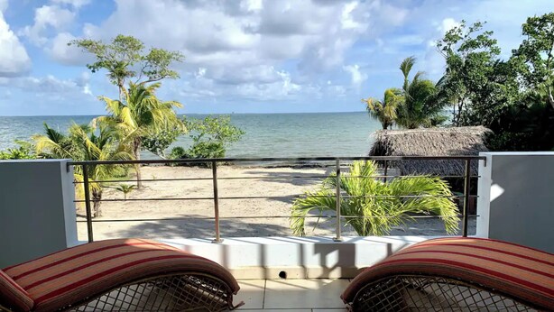 Ein Balkon mit Blick auf Strand und Meer, mit einer strohgedeckten Struktur und tropischer Bepflanzung.