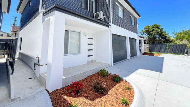 A modern two-story house with a garage, a front door, and a small garden area with red flowers.