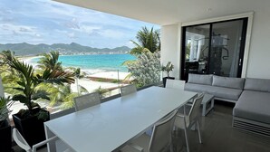 A modern living room with a large white table and chairs, a grey sofa, and a sliding glass door leading to a balcony with a view of the beach and mountains. - Beachfront Ultra Luxury - New Modern 2 Bed/2.5 Bath (Simpson Bay)