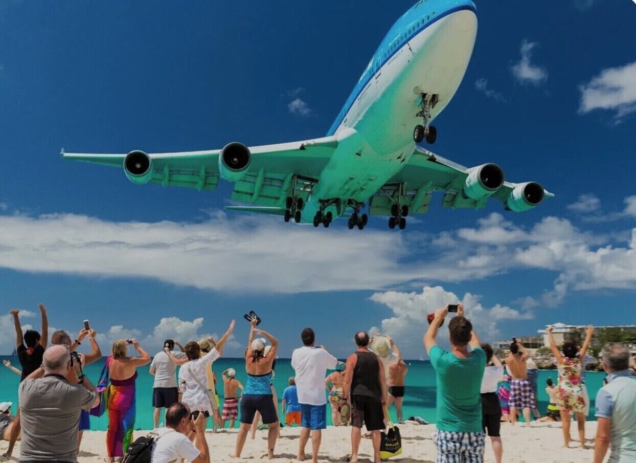Un grand avion vole à basse altitude au-dessus d’une plage, et des personnes sur le sable le regardent passer.