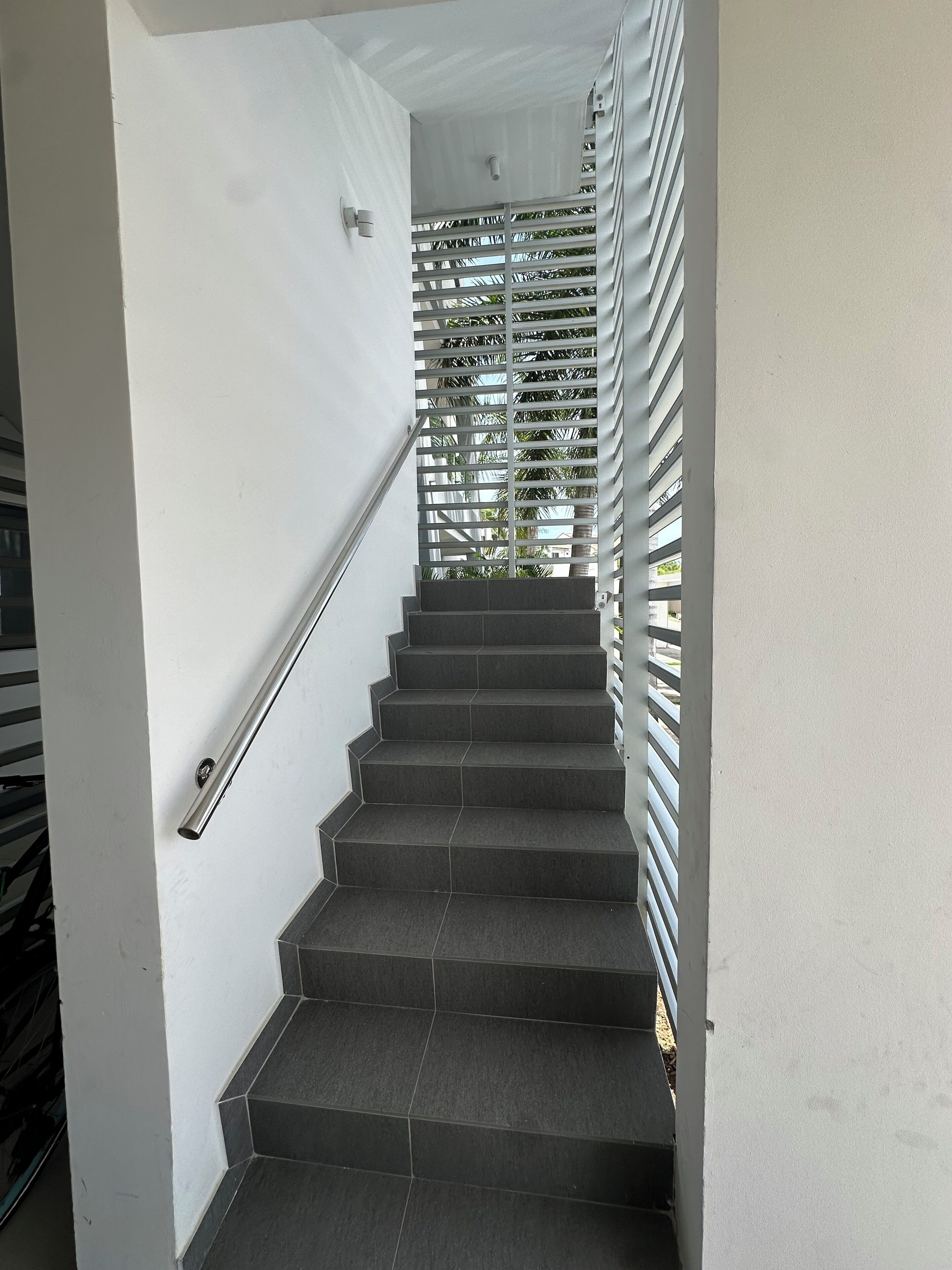A staircase with dark grey steps and a metal handrail, leading to a landing with a white wall and a security camera.