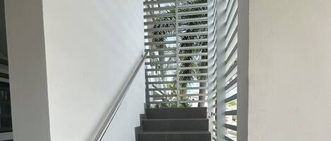 A staircase with dark grey steps and a metal handrail, leading to a landing with a white wall and a security camera.