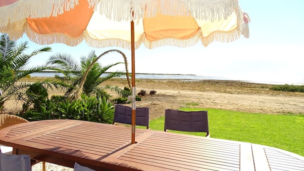 A wooden table with chairs under a large umbrella, overlooking a beach.