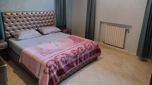 A bedroom with a tufted headboard, a bed with patterned bedding, a pink decorative throw, a nightstand, a dresser, and a radiator. - Cozy, Peaceful & Clean Studio Only 300 MAD (Ifrane)