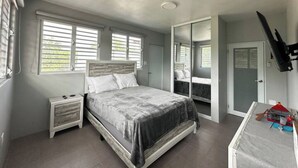 Un dormitorio con cama, mesita de noche y escritorio con silla. Hay ventanas con persianas, una puerta y un espejo.