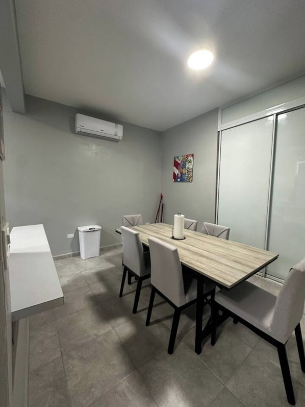 Un salón de conferencias con una mesa rectangular, seis sillas, un equipo de aire acondicionado, un cesto de basura y una pared divisoria de vidrio.