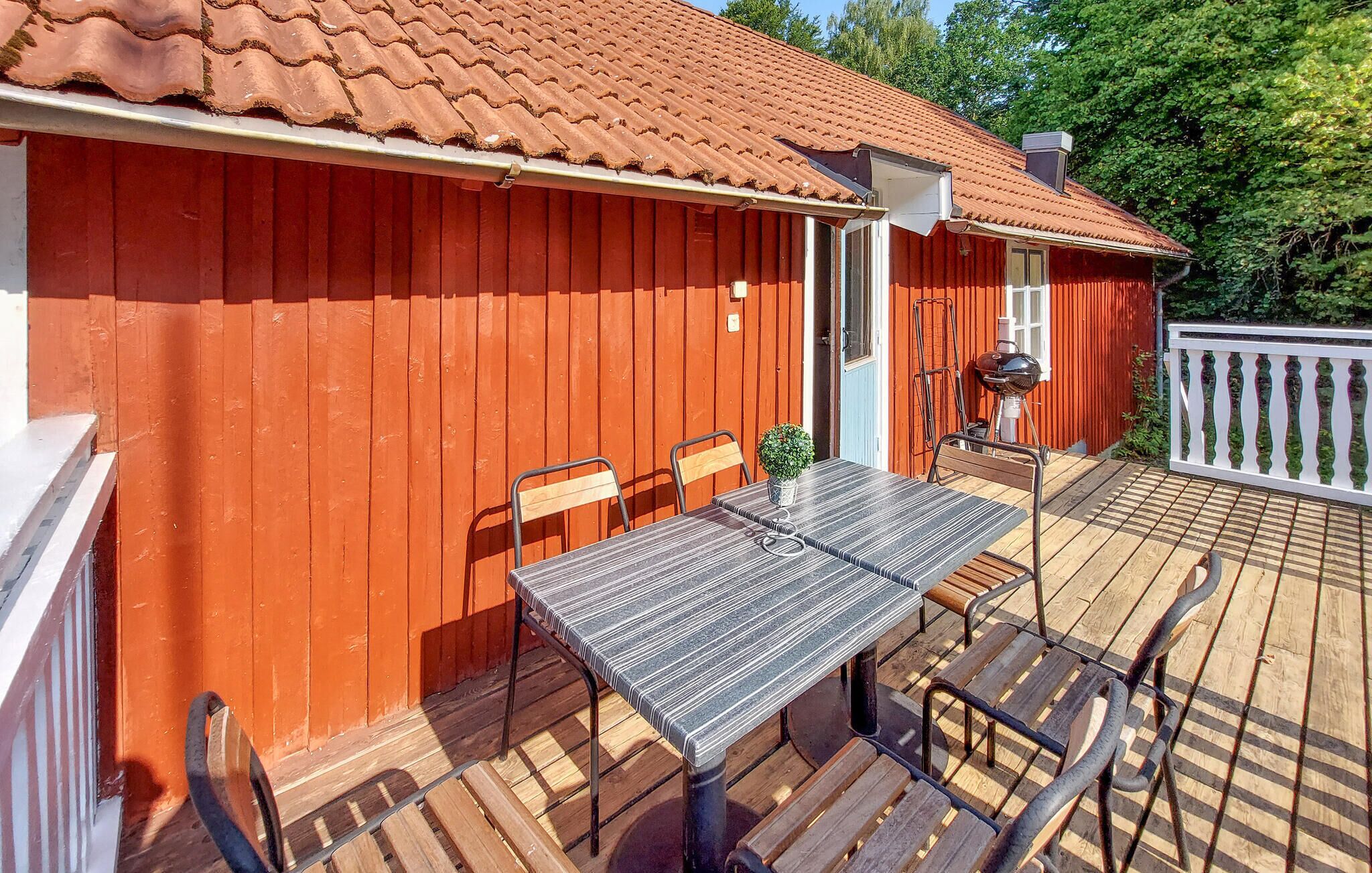 Une terrasse en bois avec une table et des chaises, un barbecue et un bâtiment rouge avec un toit en tuiles.