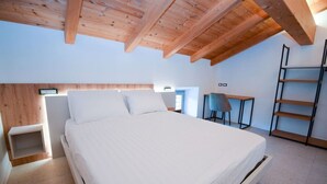 A bedroom with a wooden ceiling, a bed with white bedding, a desk with a chair, and a ladder against the wall.