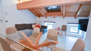 A modern living room with a glass dining table, wooden chairs, a sofa, and a TV mounted on the wall.